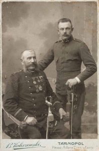 Сидить командир взводу (Zugskommandant) Імператорського і королівського австрійського військового корпусу. Він носить: Медаль за хоробрість, Срібну пам’ятну медаль за народну оборону Тиролю в 1866 році. 1908 року нагороджений Бронзовою Почесною Відзнакою За Бездоганну Службу в Корпусі Ополчення 2 ступеня за 25 років служби